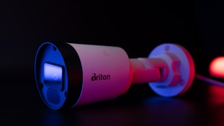 A security camera with a sleek, cylindrical design is illuminated with blue and red lighting. The brand name 'Briton' is visible on the side of the camera. The camera lens reflects a bright blue light, contrasting sharply against the dark background.