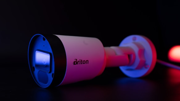 A security camera with a sleek, cylindrical design is illuminated with blue and red lighting. The brand name 'Briton' is visible on the side of the camera. The camera lens reflects a bright blue light, contrasting sharply against the dark background.