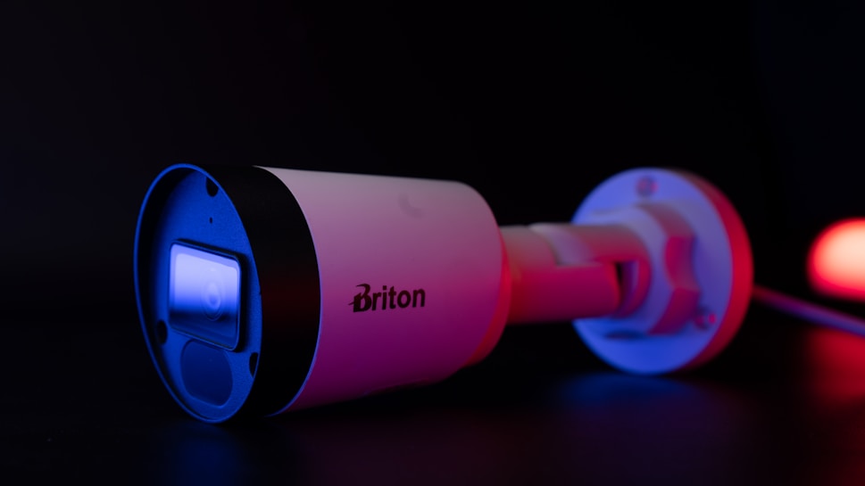 A security camera with a sleek, cylindrical design is illuminated with blue and red lighting. The brand name 'Briton' is visible on the side of the camera. The camera lens reflects a bright blue light, contrasting sharply against the dark background.