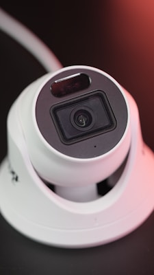 A white, dome-shaped surveillance camera with a lens in the center, positioned on a dark surface. The camera is surrounded by a subtle glow of reddish light.