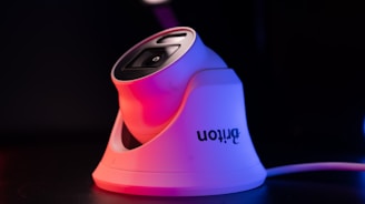 A modern white security camera with a sleek design is illuminated by contrasting red and blue lighting. The camera is placed on a dark surface, and a cord is visible extending from it.