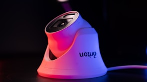 A modern white security camera with a sleek design is illuminated by contrasting red and blue lighting. The camera is placed on a dark surface, and a cord is visible extending from it.