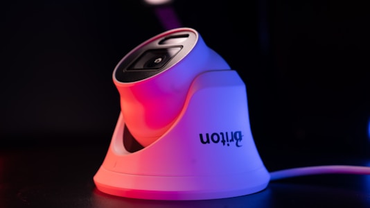 A modern white security camera with a sleek design is illuminated by contrasting red and blue lighting. The camera is placed on a dark surface, and a cord is visible extending from it.