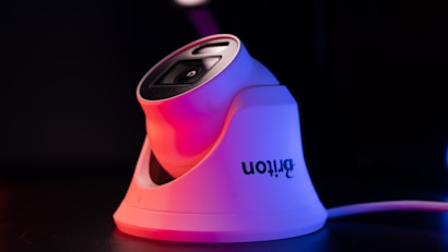A modern white security camera with a sleek design is illuminated by contrasting red and blue lighting. The camera is placed on a dark surface, and a cord is visible extending from it.