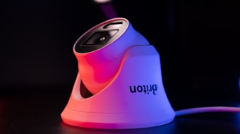 A modern white security camera with a sleek design is illuminated by contrasting red and blue lighting. The camera is placed on a dark surface, and a cord is visible extending from it.