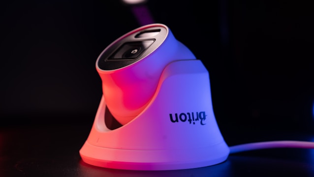 A modern white security camera with a sleek design is illuminated by contrasting red and blue lighting. The camera is placed on a dark surface, and a cord is visible extending from it.