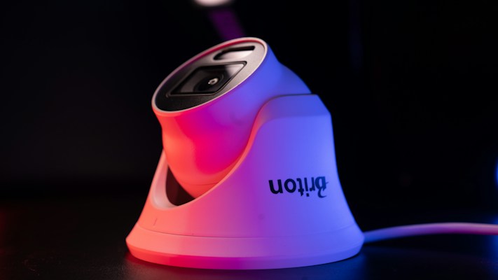 A modern white security camera with a sleek design is illuminated by contrasting red and blue lighting. The camera is placed on a dark surface, and a cord is visible extending from it.