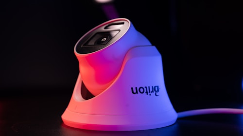 A modern white security camera with a sleek design is illuminated by contrasting red and blue lighting. The camera is placed on a dark surface, and a cord is visible extending from it.