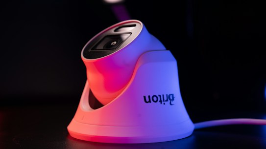 A modern white security camera with a sleek design is illuminated by contrasting red and blue lighting. The camera is placed on a dark surface, and a cord is visible extending from it.