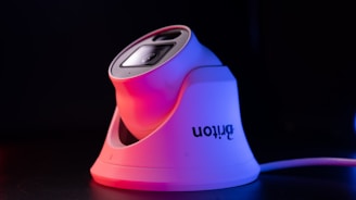 A sleek, modern security camera with a white base and lens casing, highlighted by red and purple lighting, set against a dark background. The camera brand name is visible on the side.