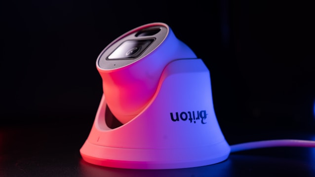 A sleek, modern security camera with a white base and lens casing, highlighted by red and purple lighting, set against a dark background. The camera brand name is visible on the side.