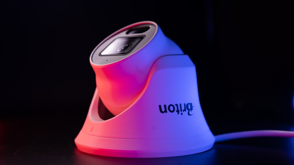 A sleek, modern security camera with a white base and lens casing, highlighted by red and purple lighting, set against a dark background. The camera brand name is visible on the side.