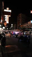 Nighttime scene on a bustling urban street featuring vibrant illuminated signage in various languages. Several tuk-tuks are lined up along the road, with a few people standing nearby. The ambient light from the signs casts colorful reflections on the street, adding to the lively atmosphere.
