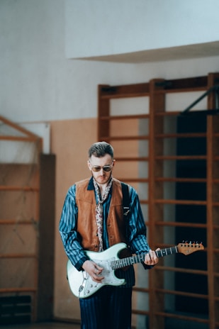 A man wearing sunglasses, a striped shirt, a brown vest, and a patterned scarf is playing an electric guitar in an indoor setting. The background consists of a wooden gymnasium structure, adding to the retro and casual vibe.