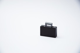 A small black toy suitcase placed against a plain white background. The suitcase appears minimalistic and features a simple handle.