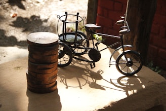 A small decorative metal bicycle is placed on a wooden surface next to a stack of wooden discs. The scene is well-lit with sunlight casting intricate shadows of the bicycle on the surface. The background features a textured brick wall and some wooden elements, suggesting an outdoor or rustic setting.