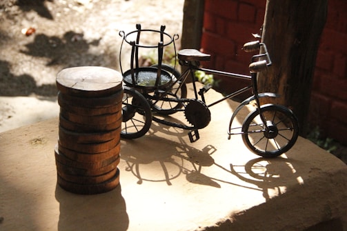 A small decorative metal bicycle is placed on a wooden surface next to a stack of wooden discs. The scene is well-lit with sunlight casting intricate shadows of the bicycle on the surface. The background features a textured brick wall and some wooden elements, suggesting an outdoor or rustic setting.