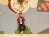 A young child stands confidently in front of a large wall mural depicting food items, including a bowl of soup with ingredients and two pieces of sushi on a platter. The child is wearing a burgundy jacket, white shirt, black pants, and red-and-white sneakers. The mural includes colors like red, green, and yellow, contributing to a playful and whimsical background.