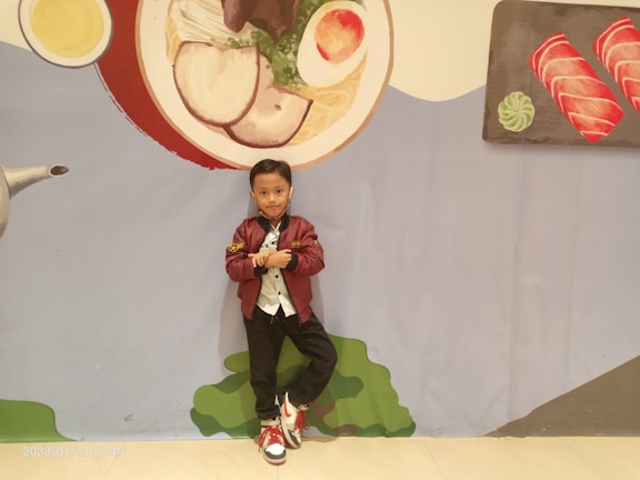 A young child stands confidently in front of a large wall mural depicting food items, including a bowl of soup with ingredients and two pieces of sushi on a platter. The child is wearing a burgundy jacket, white shirt, black pants, and red-and-white sneakers. The mural includes colors like red, green, and yellow, contributing to a playful and whimsical background.