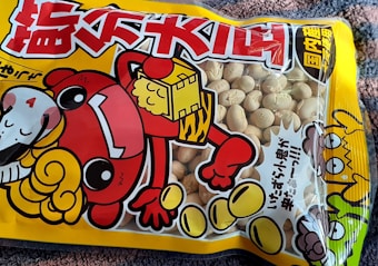 A colorful snack packaging featuring a cartoon character with a red outfit and large eyes. The character has one arm raised, holding a bag, with a sheep-like figure next to it. The packaging displays a mix of Japanese characters in bold red and black colors. The transparent section shows a collection of beige snacks inside.