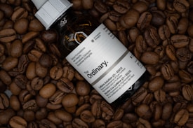A bottle labeled 'The Ordinary' lies amidst a pile of roasted coffee beans. The bottle features printed text, and light reflects off its surface, creating a shiny appearance.
