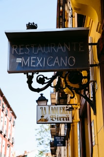 A street view showcases signs for a Mexican restaurant and a taqueria against a backdrop of colorful urban architecture. The main sign for 'Restaurante Mexicano' with vintage-style lettering is prominently displayed. Below it, another sign reads 'La Taqueria de Birra' adorned with traditional imagery. Surrounding buildings feature vibrant colors and intricate details, hinting at a lively, cultural atmosphere.