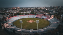 A large, mostly empty stadium viewed from above, with a cricket field in the center. The stadium is surrounded by a cityscape with buildings and greenery extending towards the horizon. The stands are circular and there are several floodlights around the field. The sky above is overcast, casting a muted light over the scene.