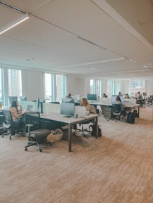 An office space with several people working at open plan desks. The room is well-lit with large windows allowing natural light to enter. Each workstation is equipped with computer monitors, chairs, and personal items. The environment appears organized and professional.
