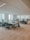 An office space with several people working at open plan desks. The room is well-lit with large windows allowing natural light to enter. Each workstation is equipped with computer monitors, chairs, and personal items. The environment appears organized and professional.
