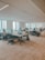 An office space with several people working at open plan desks. The room is well-lit with large windows allowing natural light to enter. Each workstation is equipped with computer monitors, chairs, and personal items. The environment appears organized and professional.