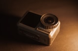 A compact action camera positioned on a dark surface, with a prominent lens on the right side and the brand name Osmo Action visible. The image is warmly lit, creating a contrast between the camera and the background.