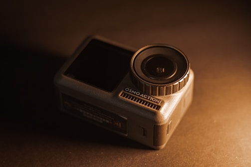 A compact action camera positioned on a dark surface, with a prominent lens on the right side and the brand name Osmo Action visible. The image is warmly lit, creating a contrast between the camera and the background.