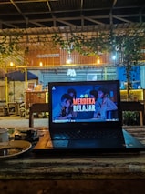 A cozy café setting with a laptop placed on a wooden table. The laptop screen displays the text 'Merdeka Belajar' with a background image of people. There is a warm, dim lighting with hanging bulbs and greenery draping from the roof. In the background, wooden furniture and a partly visible counter area can be seen.