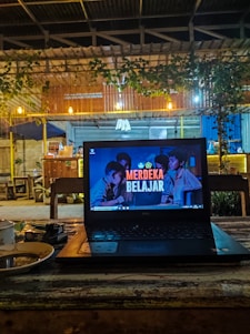 A cozy café setting with a laptop placed on a wooden table. The laptop screen displays the text 'Merdeka Belajar' with a background image of people. There is a warm, dim lighting with hanging bulbs and greenery draping from the roof. In the background, wooden furniture and a partly visible counter area can be seen.