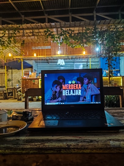 A cozy café setting with a laptop placed on a wooden table. The laptop screen displays the text 'Merdeka Belajar' with a background image of people. There is a warm, dim lighting with hanging bulbs and greenery draping from the roof. In the background, wooden furniture and a partly visible counter area can be seen.