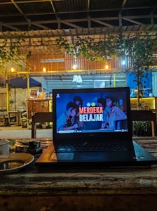 A cozy caf&eacute; setting with a laptop placed on a wooden table. The laptop screen displays the text 'Merdeka Belajar' with a background image of people. There is a warm, dim lighting with hanging bulbs and greenery draping from the roof. In the background, wooden furniture and a partly visible counter area can be seen.