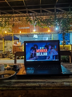A cozy caf&eacute; setting with a laptop placed on a wooden table. The laptop screen displays the text 'Merdeka Belajar' with a background image of people. There is a warm, dim lighting with hanging bulbs and greenery draping from the roof. In the background, wooden furniture and a partly visible counter area can be seen.