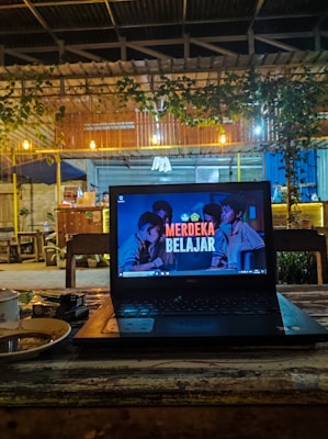 A cozy café setting with a laptop placed on a wooden table. The laptop screen displays the text 'Merdeka Belajar' with a background image of people. There is a warm, dim lighting with hanging bulbs and greenery draping from the roof. In the background, wooden furniture and a partly visible counter area can be seen.
