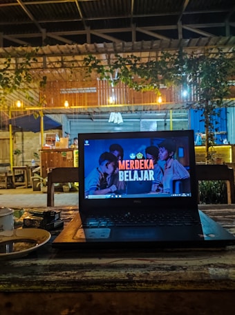 A cozy caf&eacute; setting with a laptop placed on a wooden table. The laptop screen displays the text 'Merdeka Belajar' with a background image of people. There is a warm, dim lighting with hanging bulbs and greenery draping from the roof. In the background, wooden furniture and a partly visible counter area can be seen.