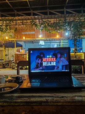 A cozy café setting with a laptop placed on a wooden table. The laptop screen displays the text 'Merdeka Belajar' with a background image of people. There is a warm, dim lighting with hanging bulbs and greenery draping from the roof. In the background, wooden furniture and a partly visible counter area can be seen.