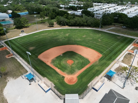 Aerial view of a well-maintained baseball field with green outfield, brown infield, pitcher's mound, and bases visible. Surrounding the field are pathways and spectator areas with some covered seating. In the background, there are trees and what appear to be residential buildings or facilities.