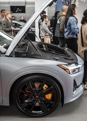 A group of people gather around a modern electric car displayed in an indoor showroom. The car's hood is open, revealing the front trunk area. The car has sleek, black wheels with a striking design, and the brake calipers are painted in bright yellow.