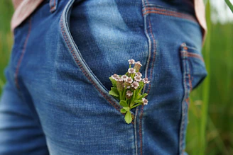 A close-up view of a person wearing blue denim jeans with a small bouquet of wildflowers tucked into the front pocket. The jeans have visible orange stitching, and the background shows a blurred outdoor setting with green foliage.