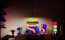 A modern gaming setup features a dual monitor arrangement with vibrant, abstract backgrounds. A glowing '2024' sign is mounted above the monitors. The desk includes a sleek gaming PC with LED accents, and the room is bathed in ambient lighting from multiple RGB light sources. Shelves with decorative items and a chair complete the workspace.