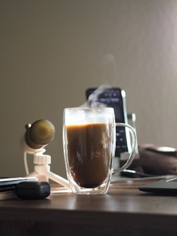 A cozy workspace scene with a glass mug of steaming coffee on a wooden desk. Nearby, there is a gold-colored microphone on a stand, an open laptop, and a smartphone propped up in the background. The overall setting suggests a relaxed atmosphere with warm lighting.