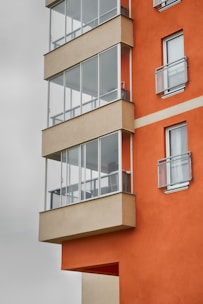 A modern apartment building with an orange and beige facade, featuring several glass-enclosed balconies. The design is geometric, with sharp angles and a clean, contemporary look.