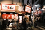 A bustling street scene at night in an urban setting with a traditional Japanese ramen shop. The shop is adorned with vibrant red signage and lanterns, with people visible inside and out, some sitting and others standing. The street is lined with various storefronts, illuminated by neon lights, creating a lively and atmospheric scene.