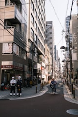 A narrow urban street flanked by tall buildings with numerous signs and advertisements. Power lines crisscross overhead. Two security personnel stand on the left side, and a person rides a bicycle down the street.