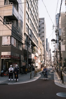 A narrow urban street flanked by tall buildings with numerous signs and advertisements. Power lines crisscross overhead. Two security personnel stand on the left side, and a person rides a bicycle down the street.