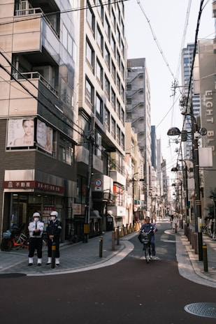 A narrow urban street flanked by tall buildings with numerous signs and advertisements. Power lines crisscross overhead. Two security personnel stand on the left side, and a person rides a bicycle down the street.